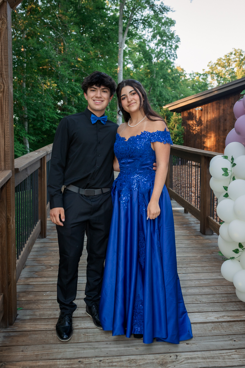 A young couple in formal wear posing on a wooden deck outdoors; woman in a blue sequined off‑the‑shoulder gown, man in black with a blue bow tie.