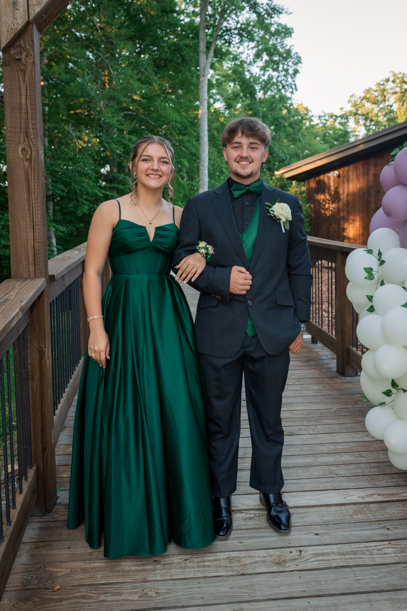 Young couple in formal attire posing arm-in-arm on a wooden deck with green outfits and a balloon display nearby.
