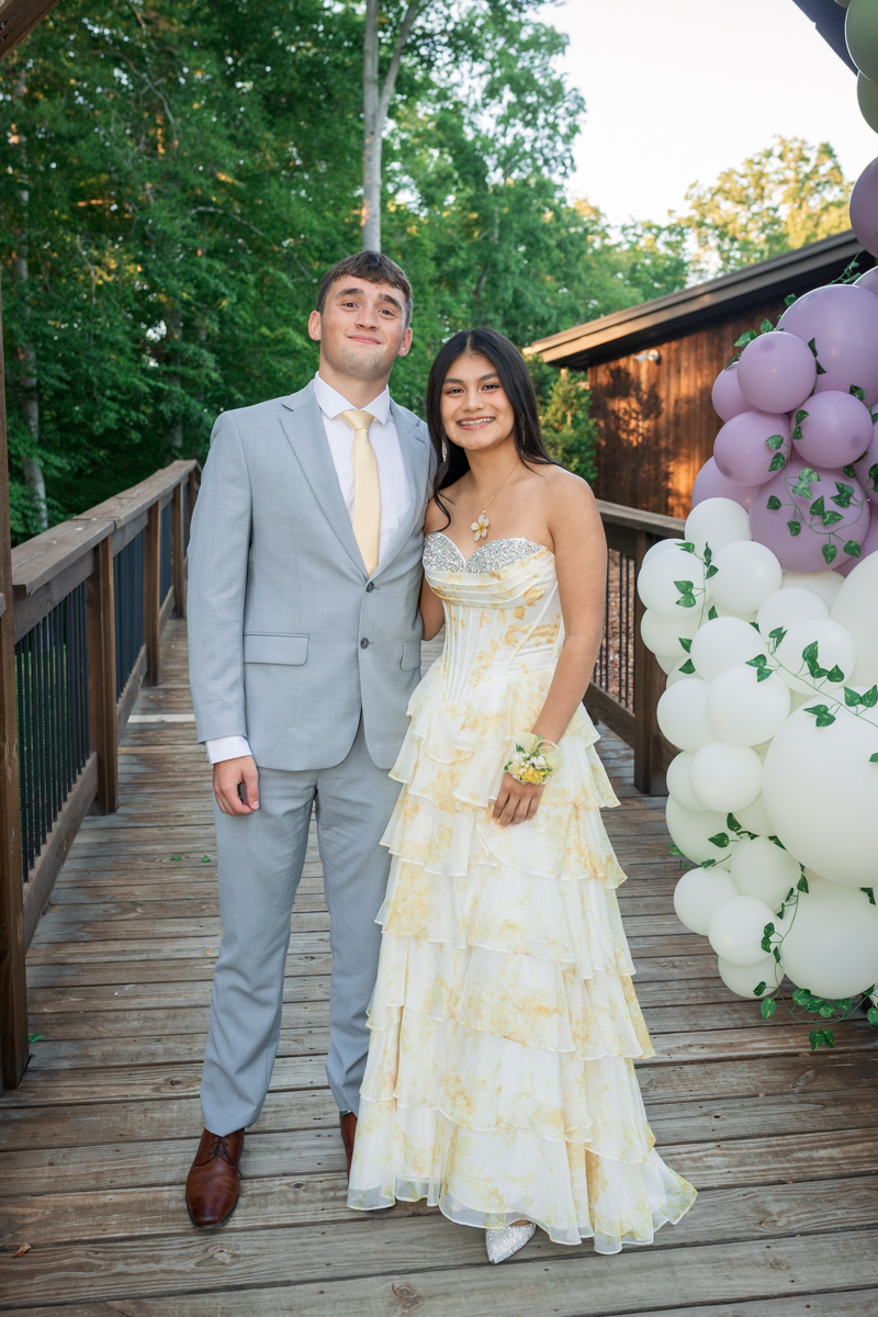 Smiling couple posing on a wooden deck: man in a light gray suit with a cream tie and brown shoes, woman in a yellow-and-white strapless gown with a floral bracelet, beside a balloon arch.