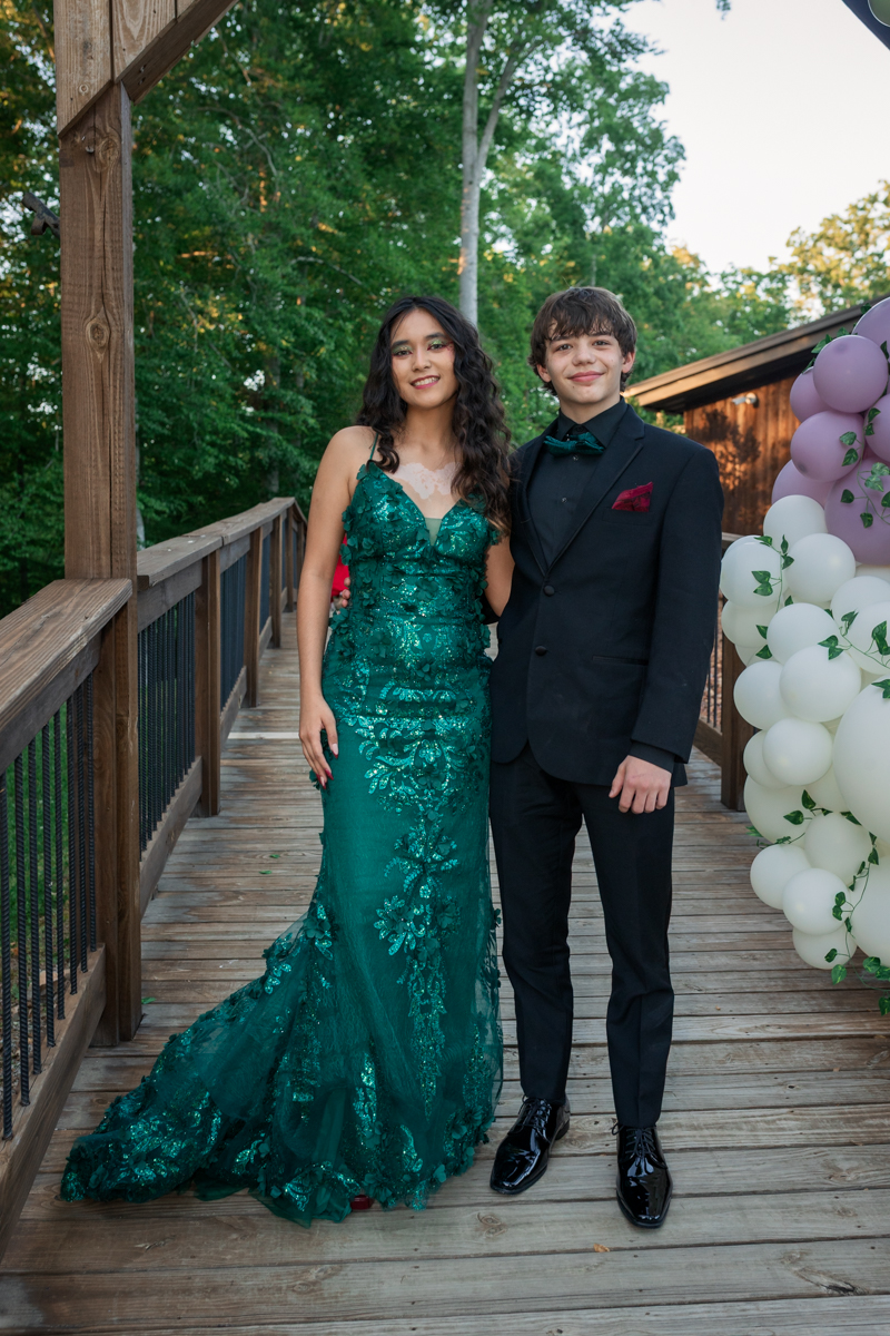 Couple in formal wear pose on a wooden bridge outdoors, woman in a green lace gown and man in a black suit with a bow tie, balloons nearby.