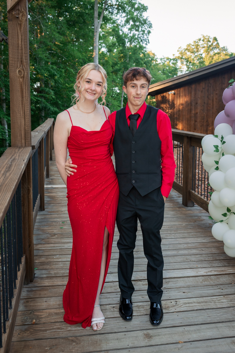 Two teens in formal attire posing on a wooden walkway; girl in a red sparkly gown and slit, boy in a red shirt with a black vest and tie, smiling together on a deck.