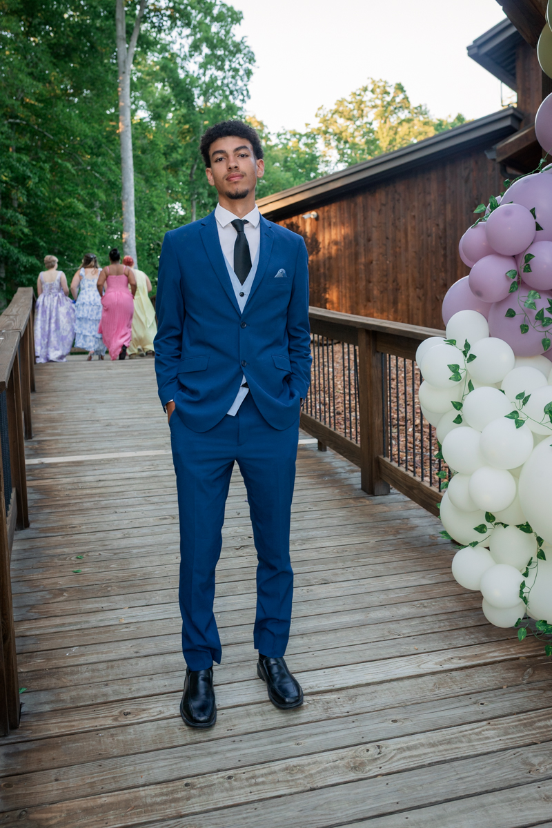 Young man in a blue suit posing on a wooden deck at an outdoor celebration with pastel balloons nearby.