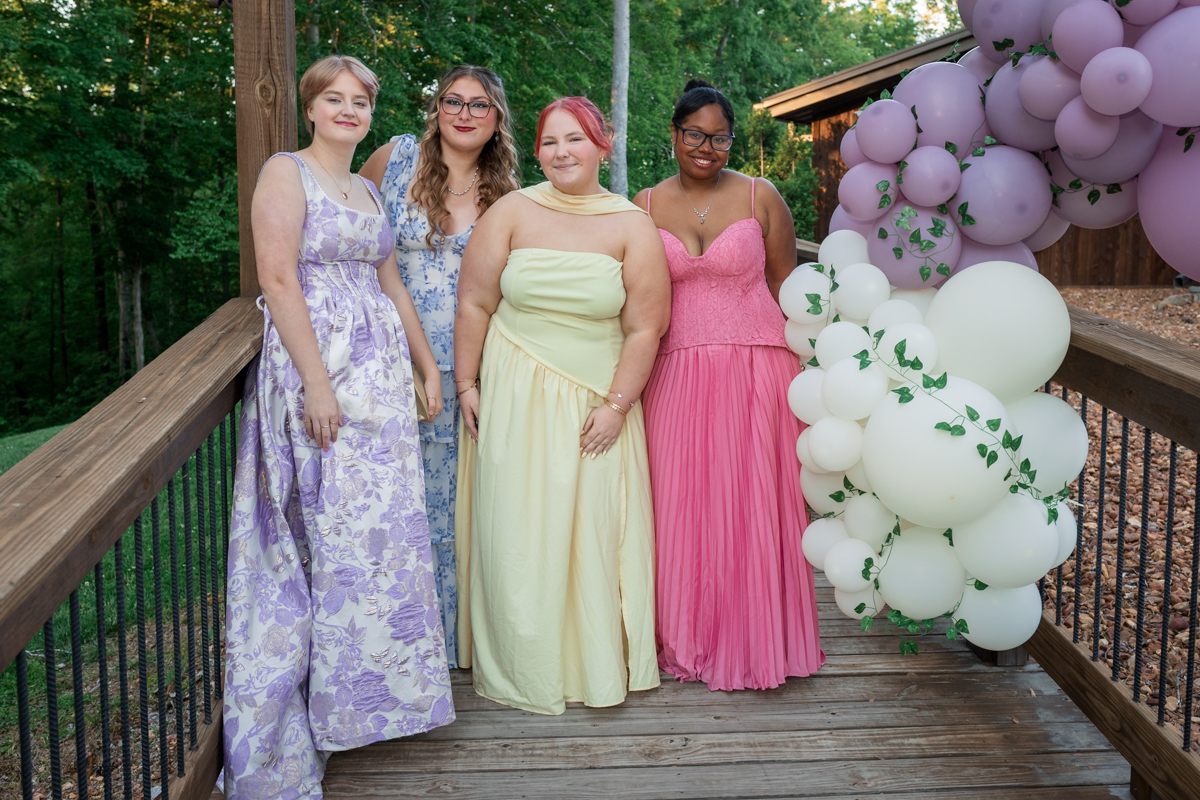 Five young women in pastel formal dresses pose on a wooden deck with lavender and pink balloons beside them.