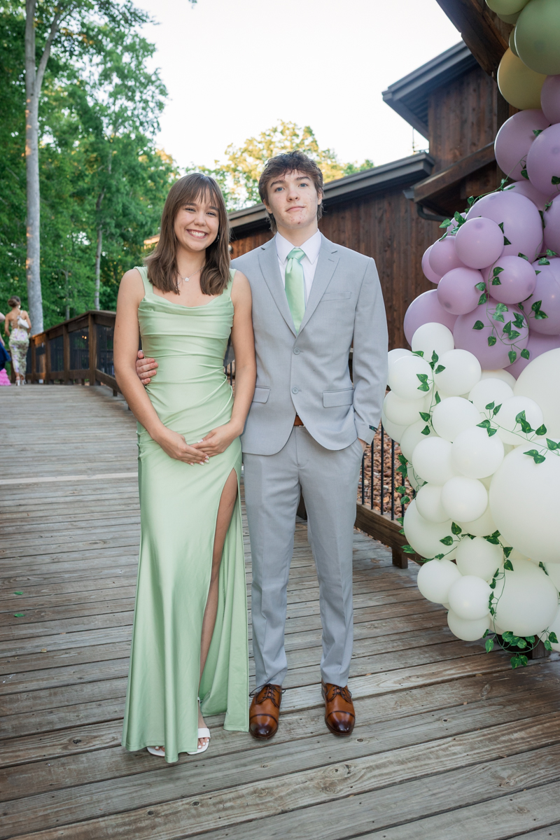 Smiling couple in mint dress and light gray suit posing on a wooden deck beside a pastel balloon arch at an outdoor event