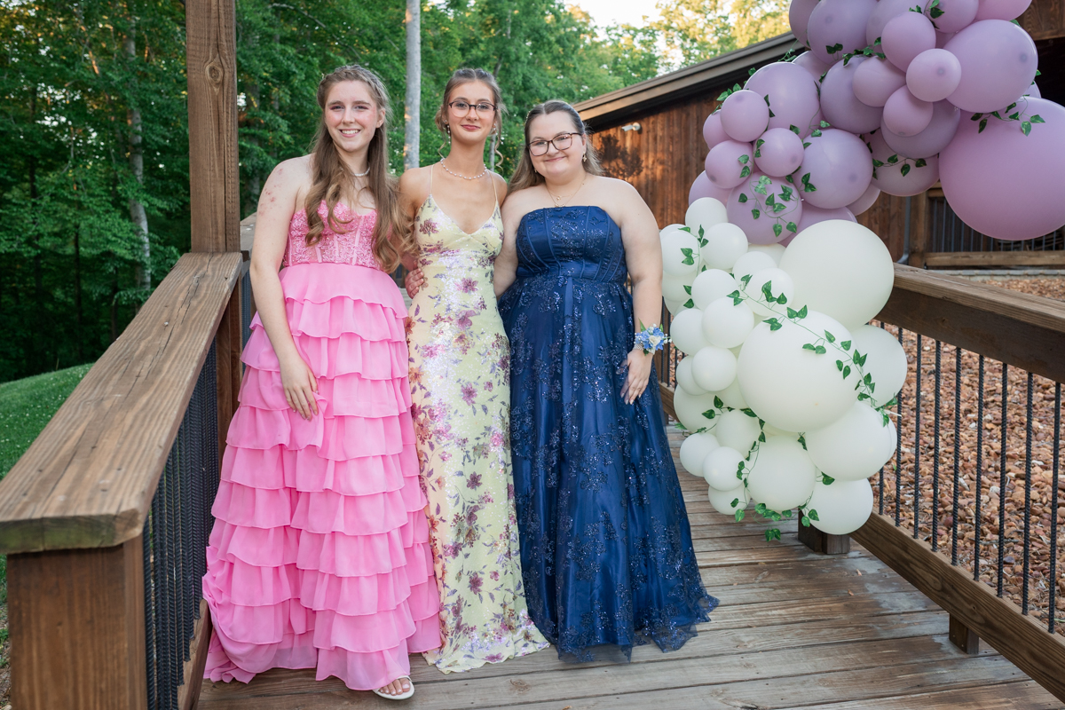 Three young women in formal gowns stand together on a wooden deck with pastel balloon decorations to the right, smiling at the camera.