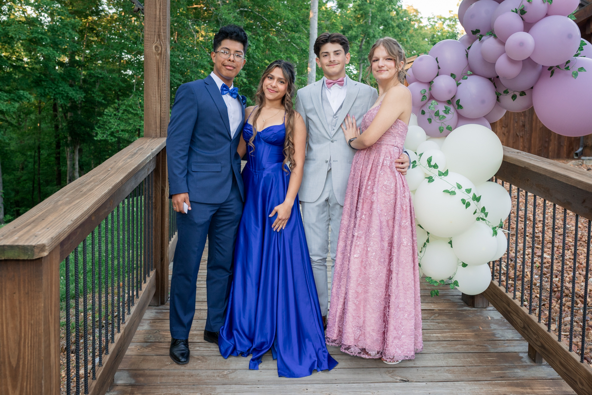 Four teens in formal wear pose on a wooden deck with a lavender and white balloon arch behind them outdoors, likely at prom or a party.