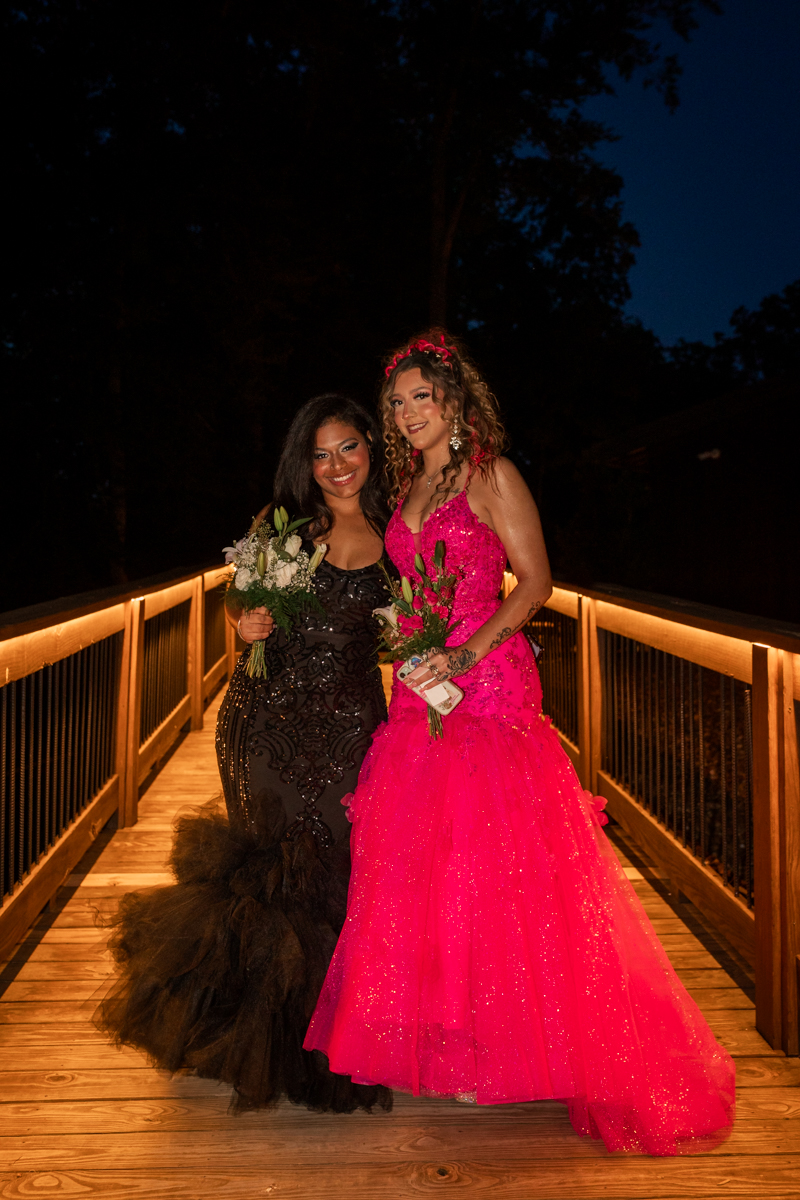 Two women in formal gowns pose on a lit wooden bridge at night, holding bouquets steadied beside them.
