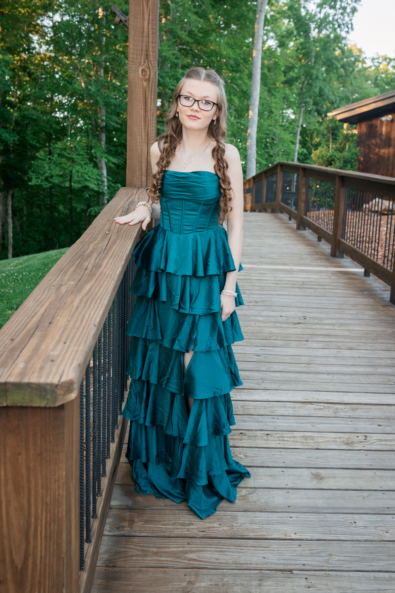 Young woman in a teal strapless, tiered gown stands on a wooden deck outdoors.