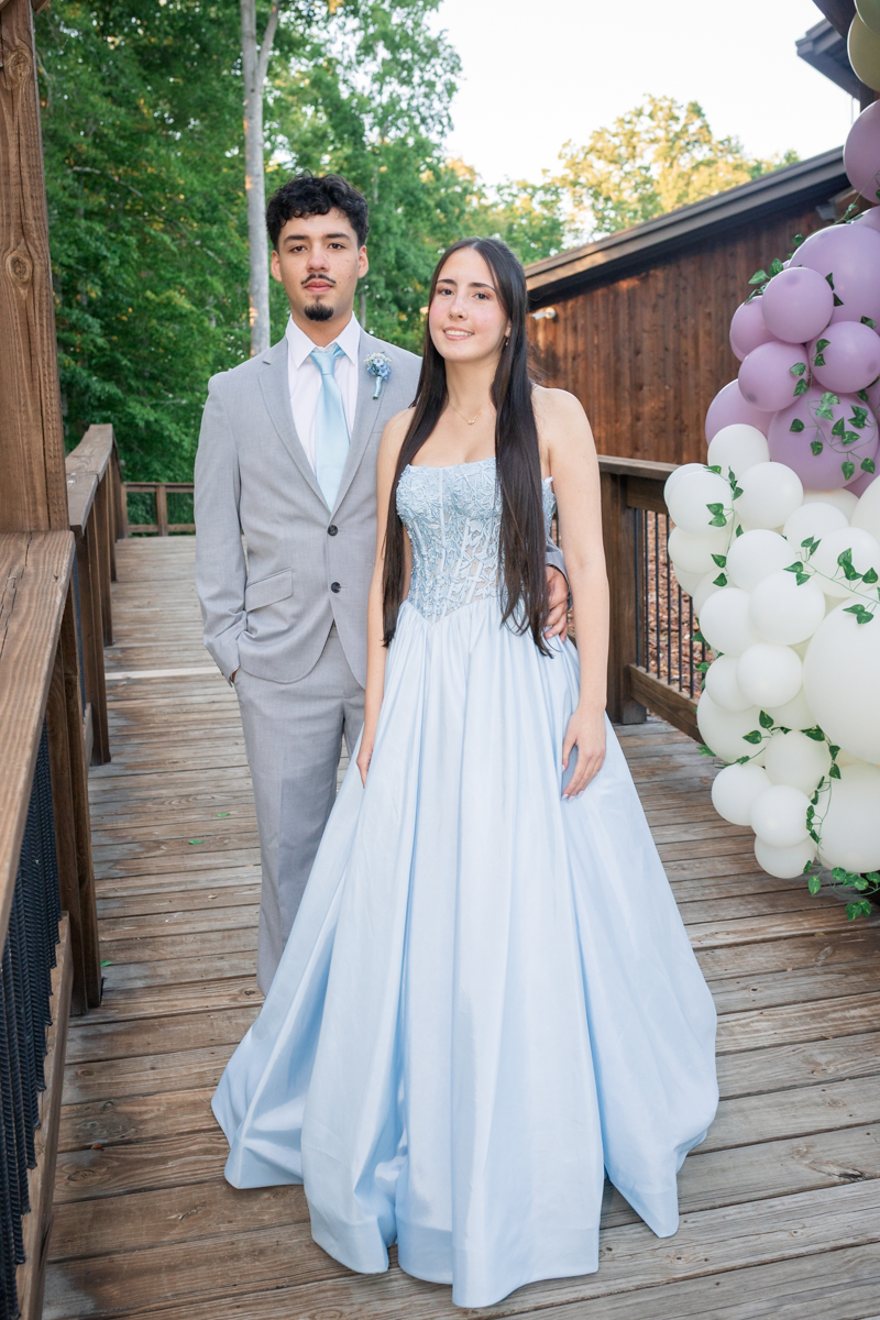 Couple in formal attire posing on a wooden deck outdoors, woman in a light blue ball gown and man in a gray suit, with pastel balloons on the right side.