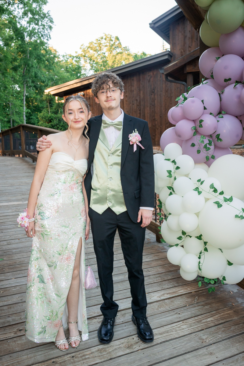A couple dressed in formal attire stands on a wooden deck beside a pastel balloon arch, posing for a photo outside.
