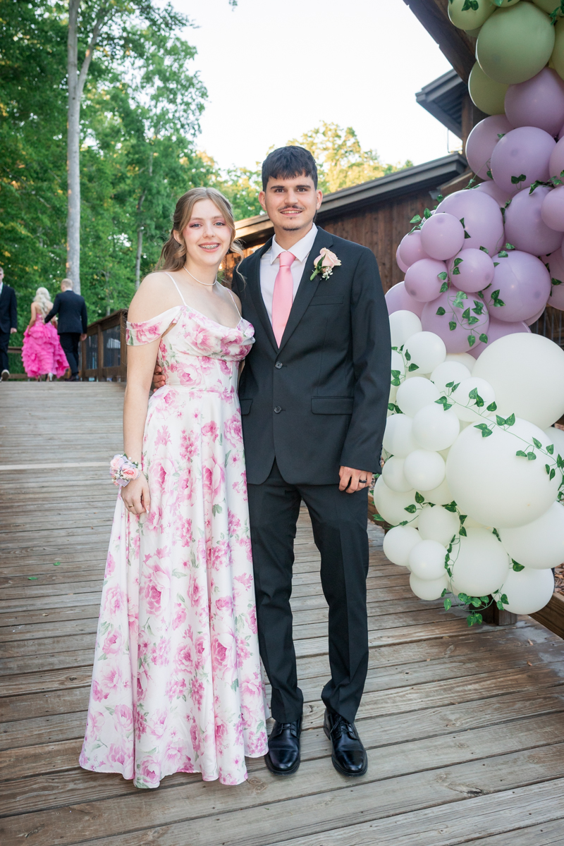 Couple posing outdoors at a formal event; woman in a pink floral gown with a corsage and man in a black suit with a pink tie, beside a pastel balloon arch.