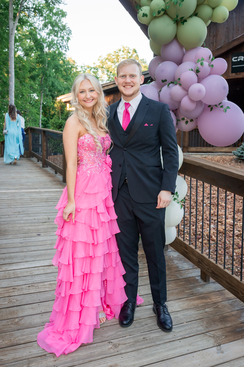 Couple in formal attire posing on a wooden deck with a pastel balloon arch; woman in a pink ruffled gown, man in a dark suit with a pink tie.