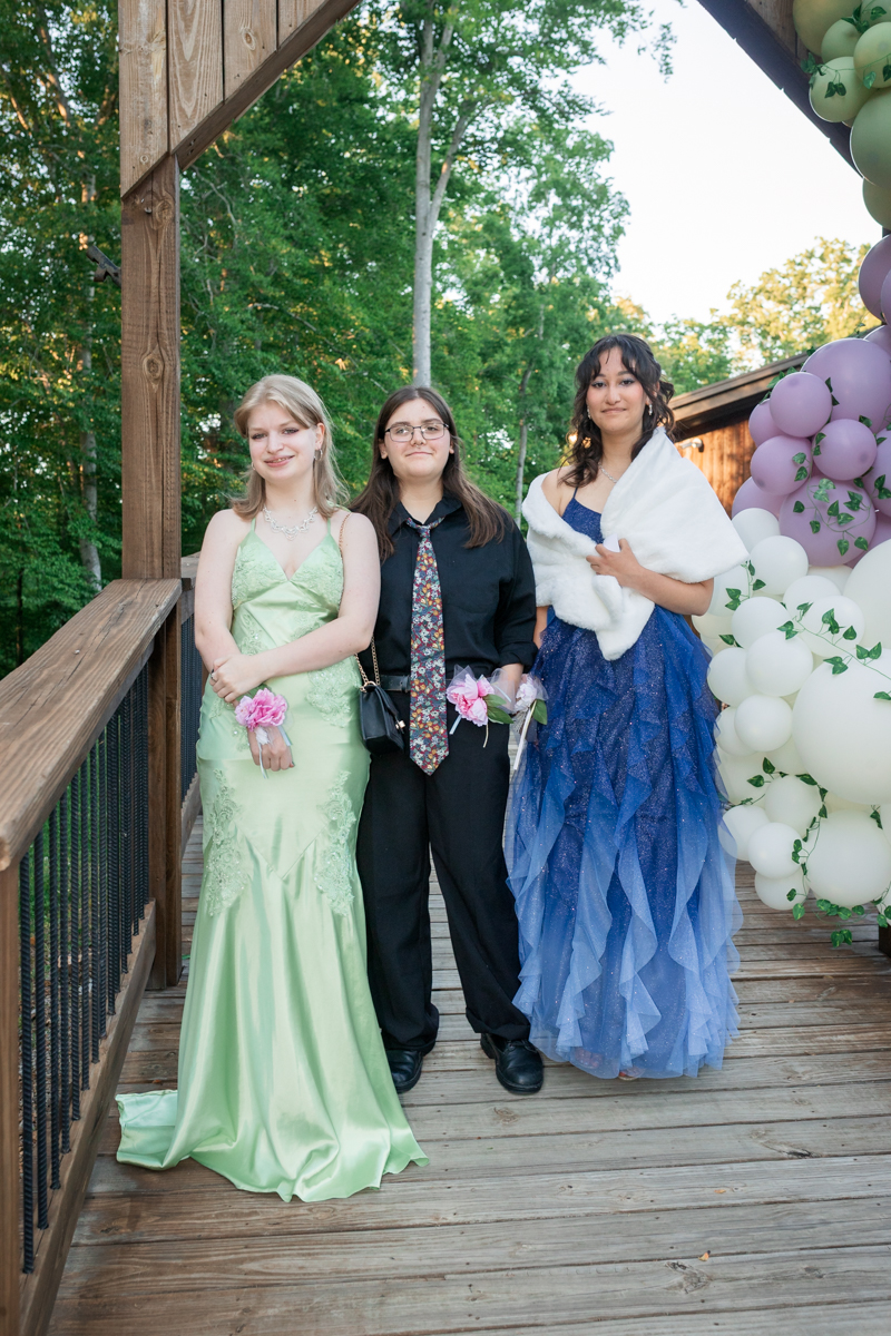 Three teens in formal attire pose on a wooden deck with pastel balloons at an outdoor celebration.