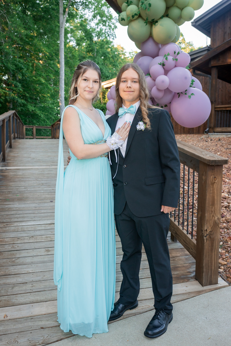 Couple in formal wear posing outdoors, girl in a light blue gown and gloves, boy in a black suit with a turquoise bow tie, pastel balloons behind them.