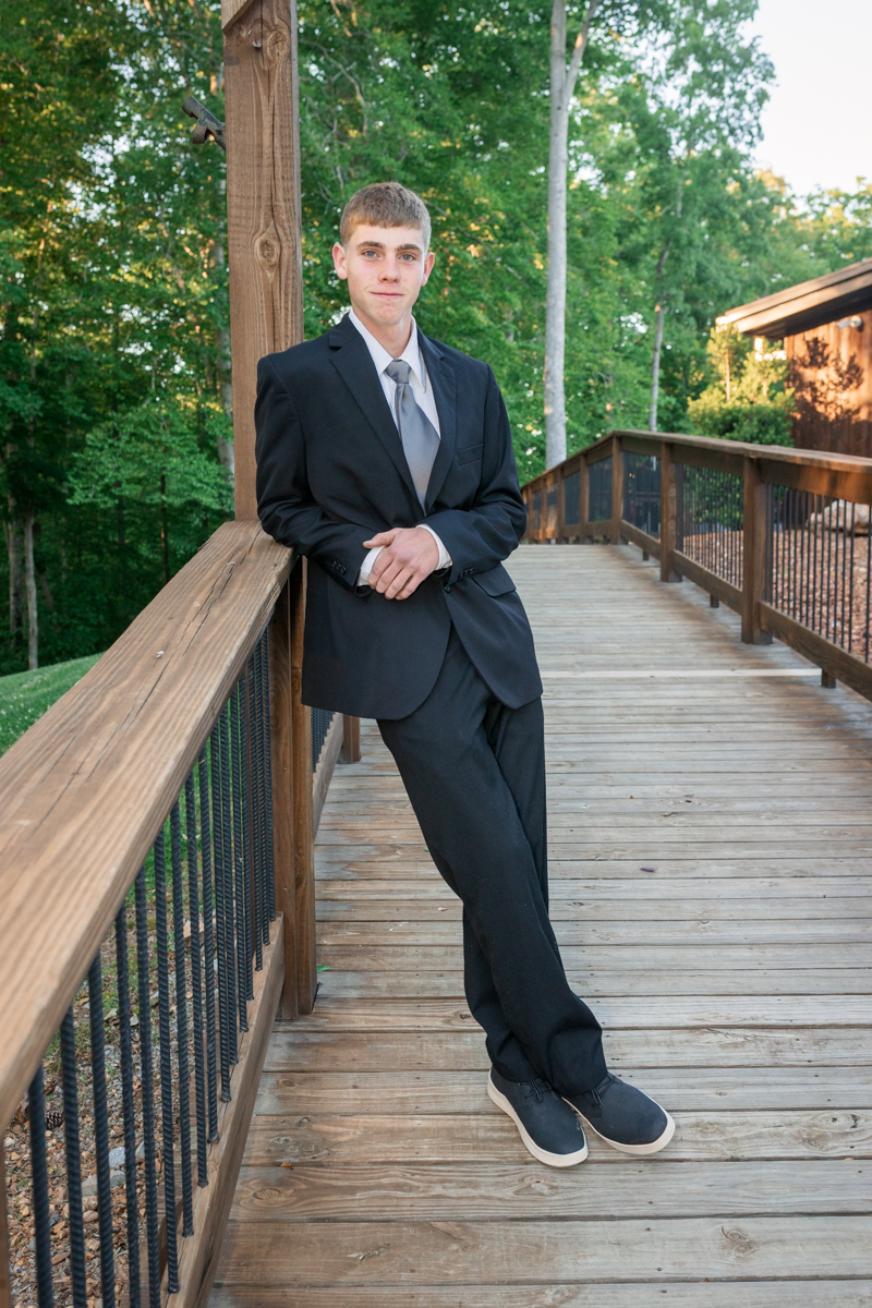 Young man in a black suit and gray tie leaning against a wooden railing on an outdoor deck with trees in the background.