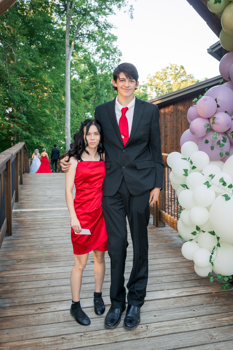 Two teens in formal wear pose on a wooden bridge beside a pastel balloon arch; girl in red dress, boy in black suit.