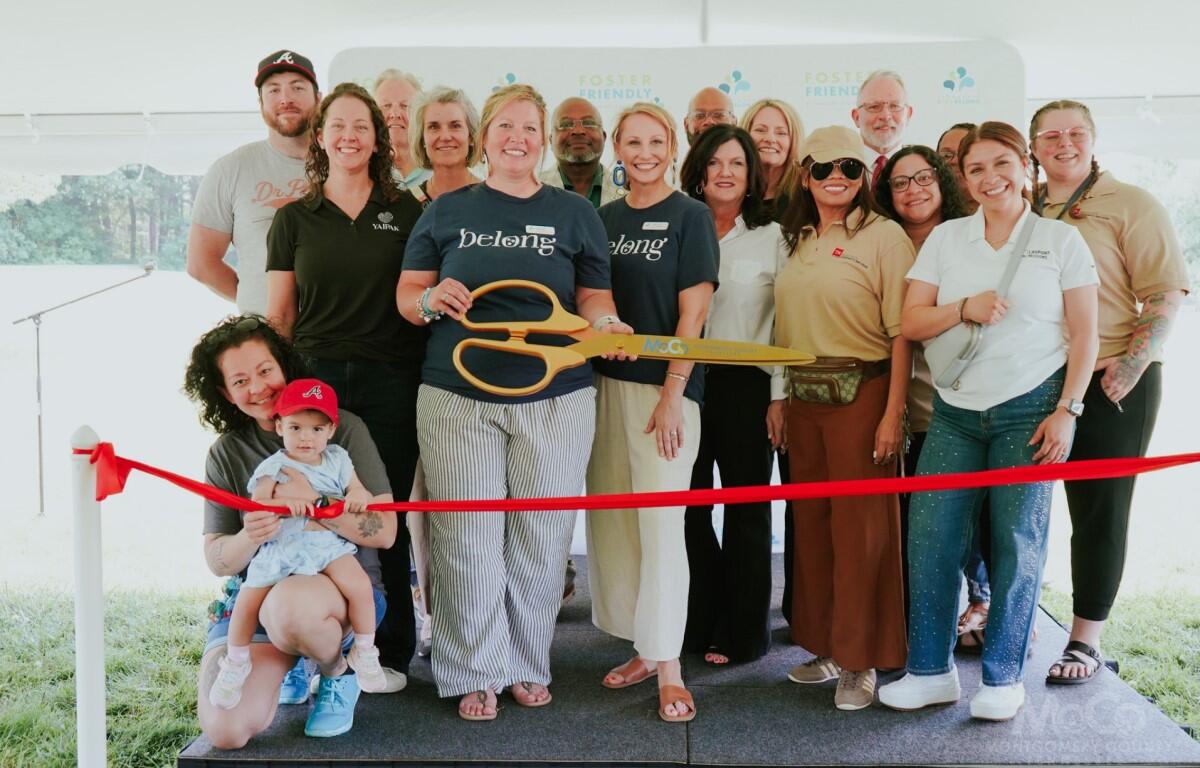 Group of diverse adults posing with oversized orange scissors at a ribbon-cutting ceremony under a white tent with a red ribbon in the foreground.