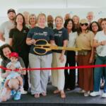 Group of diverse adults posing with oversized orange scissors at a ribbon-cutting ceremony under a white tent with a red ribbon in the foreground.