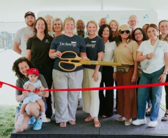 Group of diverse adults posing with oversized orange scissors at a ribbon-cutting ceremony under a white tent with a red ribbon in the foreground.