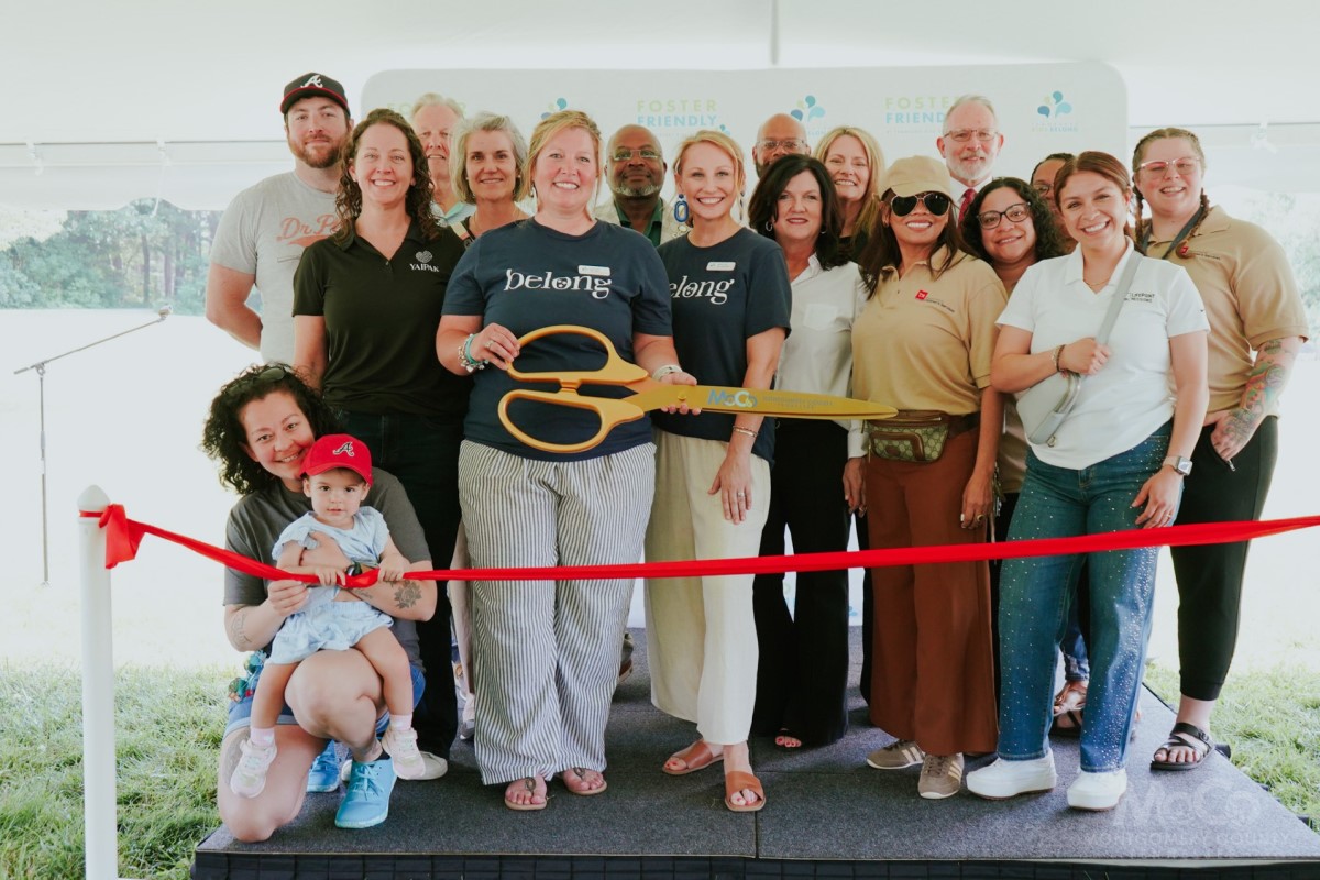 Group of diverse adults posing with oversized orange scissors at a ribbon-cutting ceremony under a white tent with a red ribbon in the foreground.