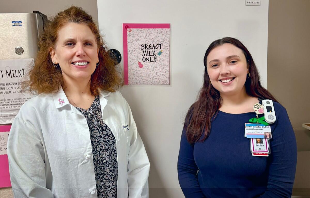 Two healthcare workers stand and smile in a clinic near a fridge with a pink 'Breast Milk Only' sign on the door.
