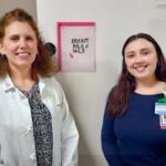 Two healthcare workers stand and smile in a clinic near a fridge with a pink 'Breast Milk Only' sign on the door.