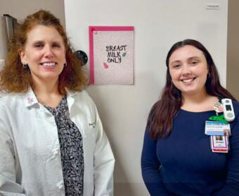 Two healthcare workers stand and smile in a clinic near a fridge with a pink 'Breast Milk Only' sign on the door.