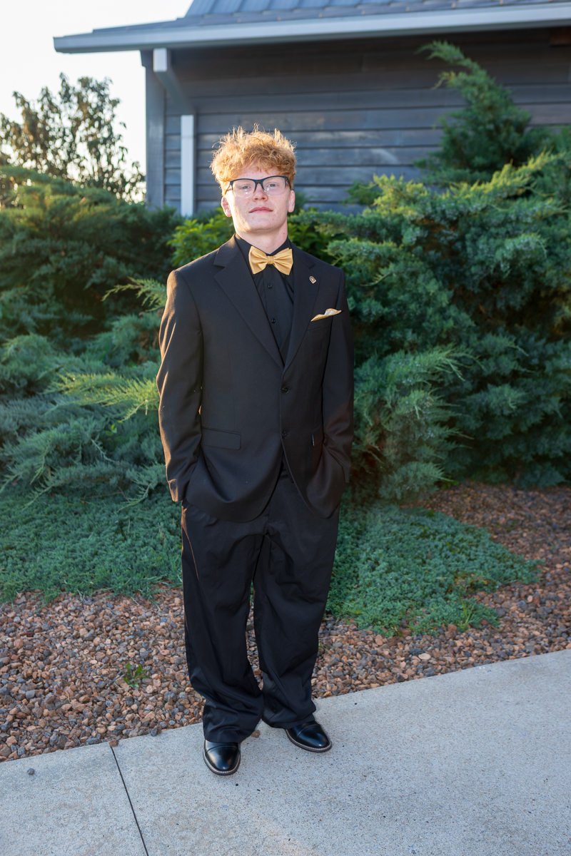 Young man in a black suit with a gold bow tie and pocket square stands with hands in pockets outdoors, near green shrubs and a wooden building.