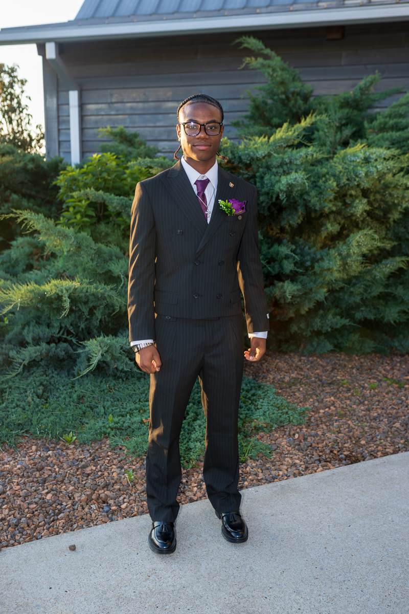 Young man in a dark pinstripe double-breasted suit with a purple boutonniere, standing outdoors by shrubs.