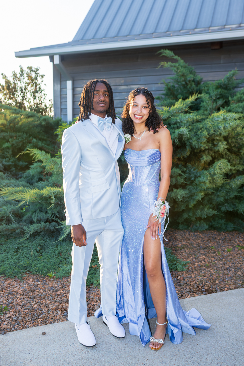 Young couple in formal attire posing outdoors, man in a light blue suit and woman in a sparkly blue gown with a leg slit.