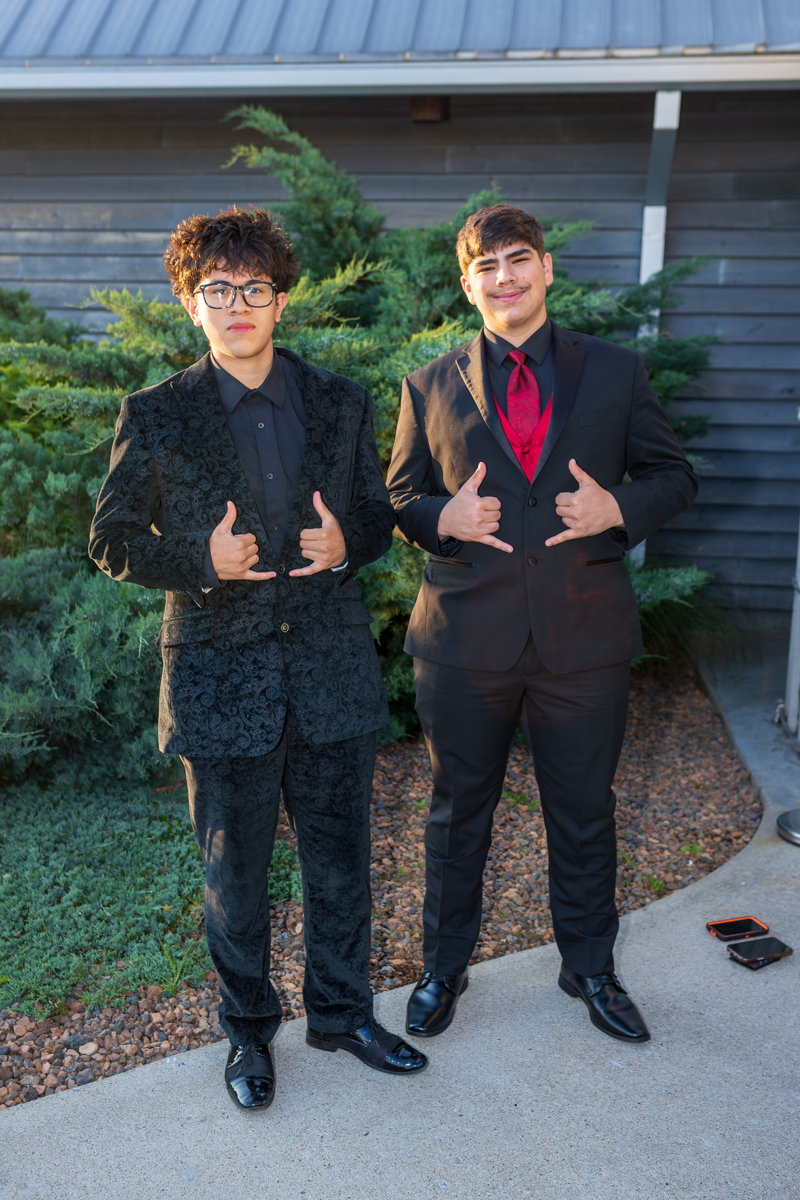Two young men in formal suits posing outdoors, both making shaka hand gestures toward the camera.