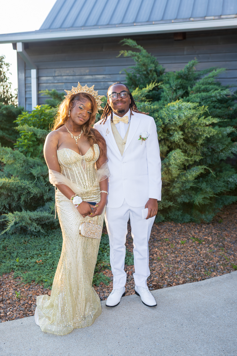 Couple in formal wedding attire posing outdoors; woman in a gold gown with a crown, man in a white suit with a gold bow tie.