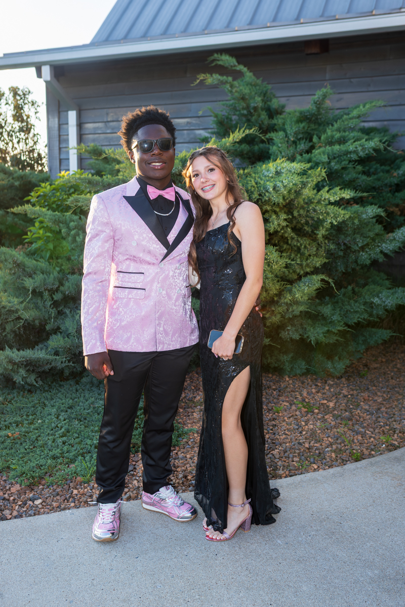 Couple posing outdoors in formal wear: young man in a pink patterned suit with a pink bowtie and sneakers, woman in a black sequined gown holding a clutch.