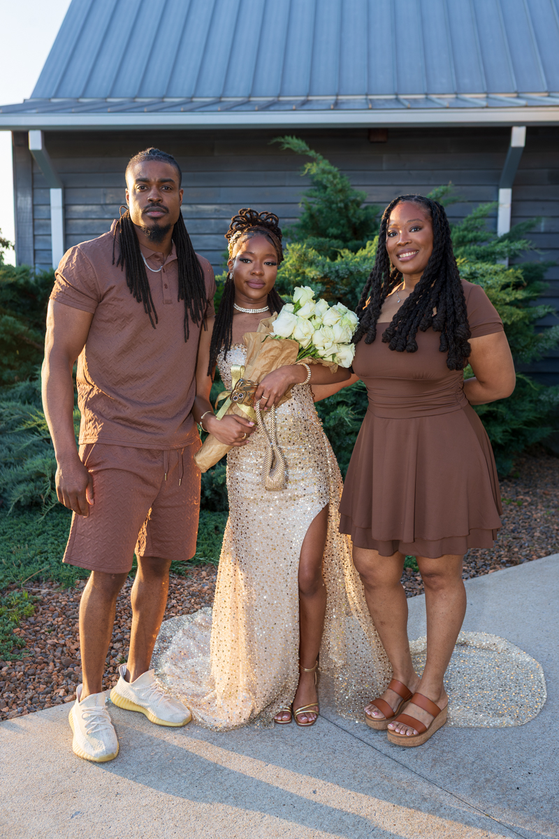 Three people posing outdoors: a man in brown outfit on the left, a center woman in a gold sequin gown holding a white bouquet, and a woman in brown on the right.