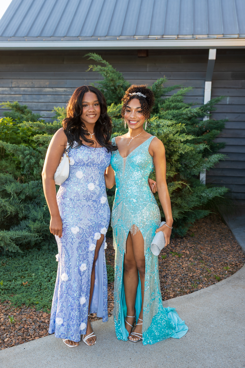 Two young women in formal gowns pose together outdoors, smiling; one in lavender lace with white embellishments, the other in turquoise sequined gown with a high slit and fringe detail.