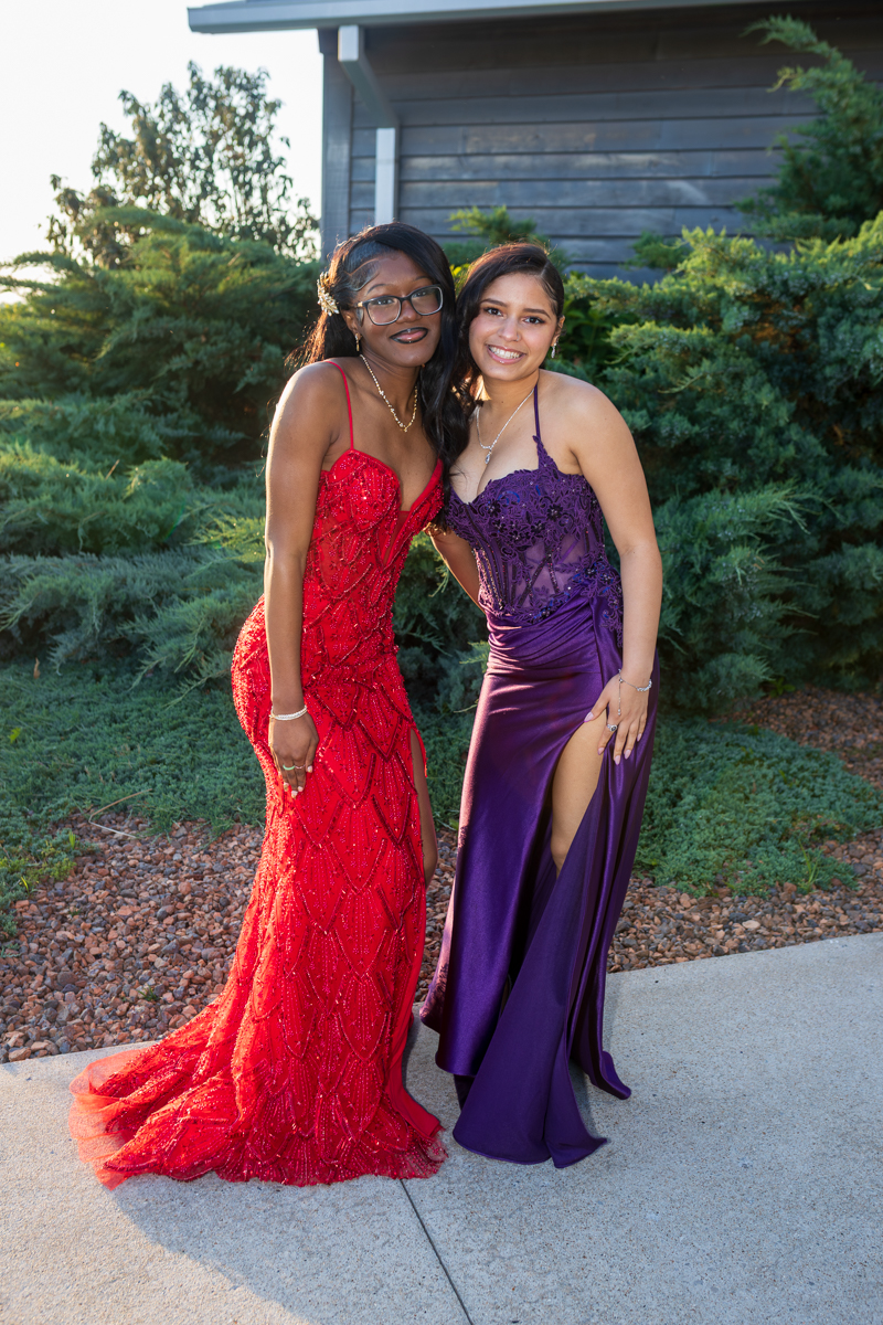 Two young women in formal gowns posing together outdoors, one in a red beaded dress and the other in a purple gown with a high slit, smiling at the camera.