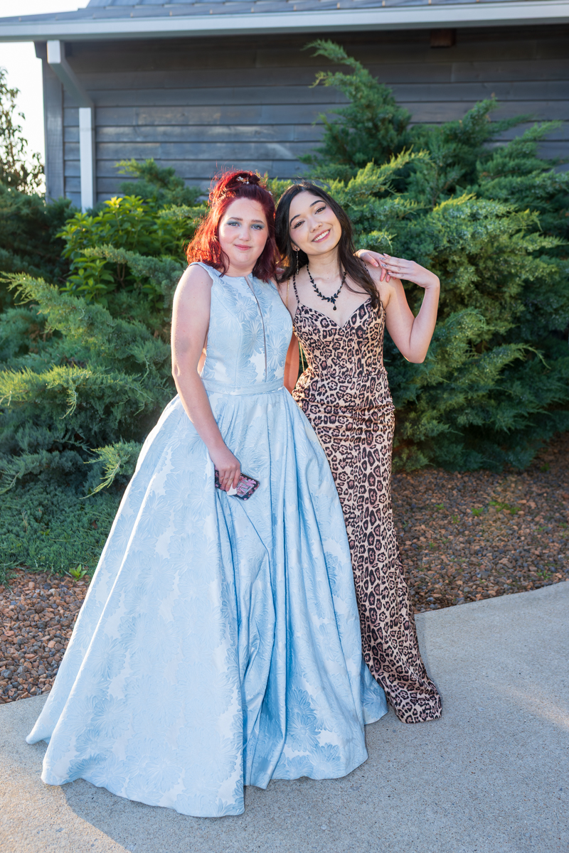 Two women in formal gowns posing together outside, one in a light blue ball gown and the other in a leopard-print dress, smiling at the camera.