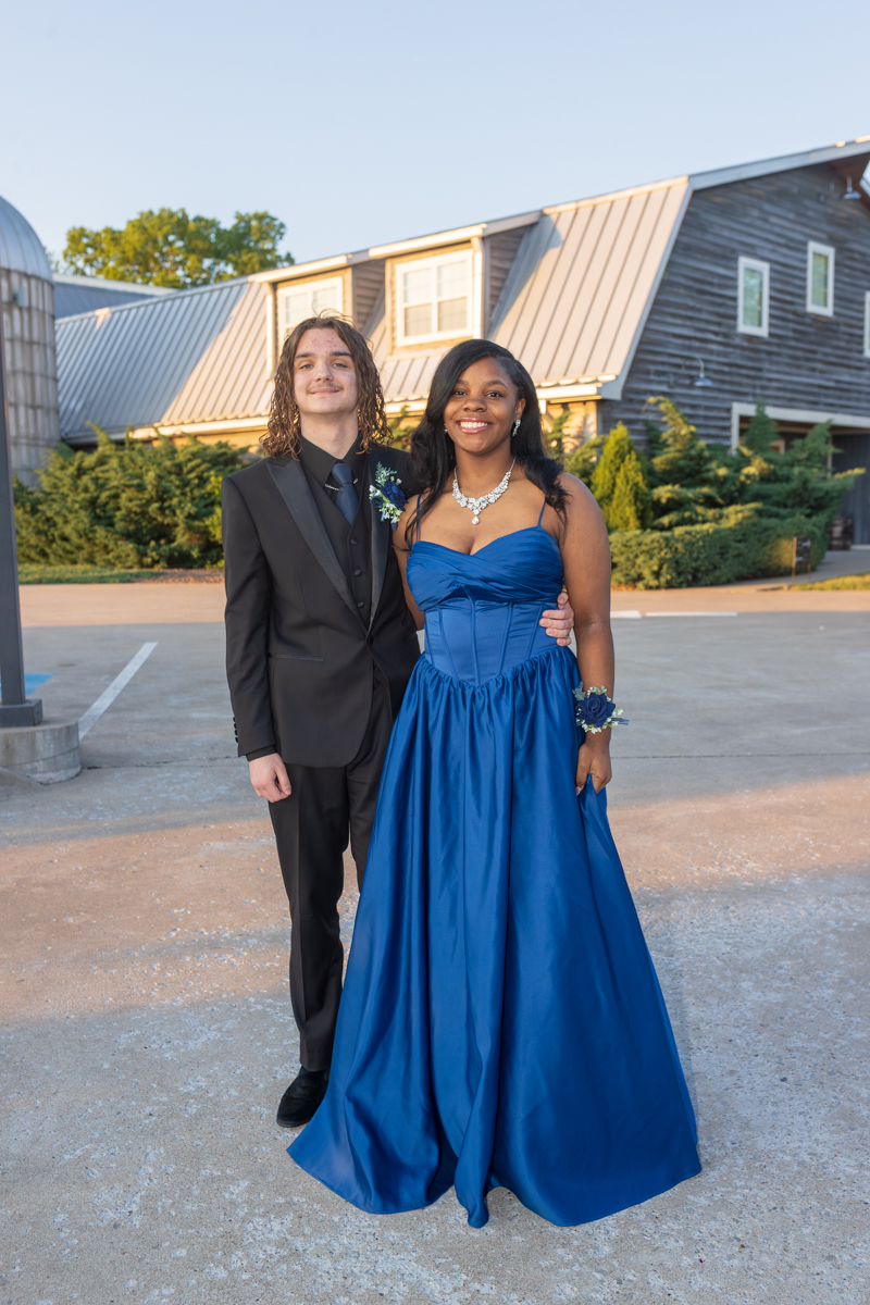 Two young people in formal attire pose arm in arm outside a modern building, the young woman in a royal blue strapless gown with jewelry and a corsage, and the young man in a black suit with a boutonniere.