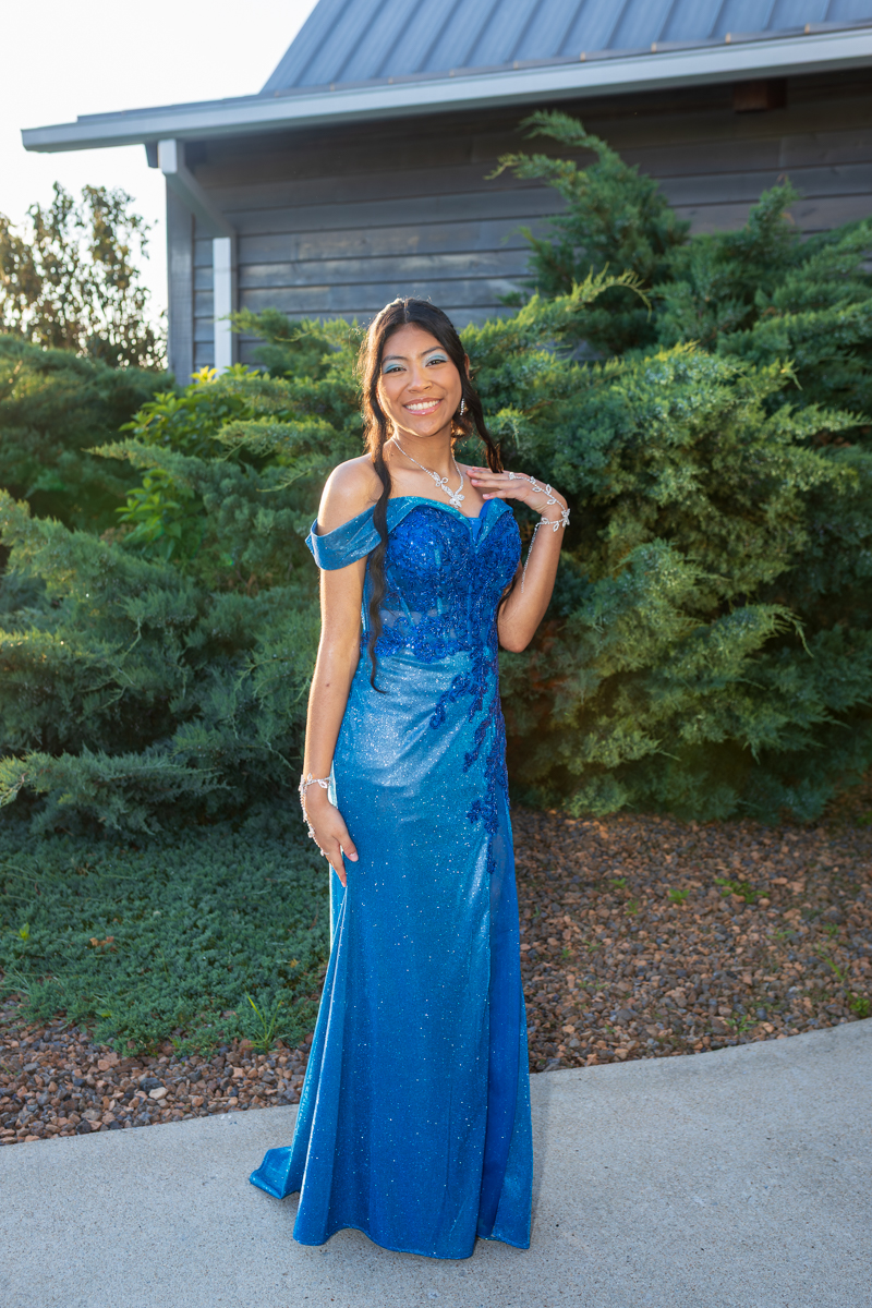 Smiling young woman in an off-the-shoulder blue evening gown with jewelry, standing outside among shrubs.