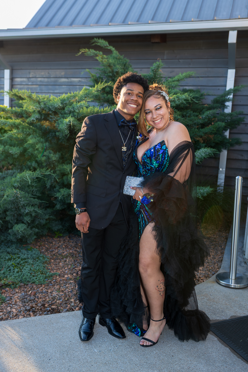 Smiling couple posing together outdoors; man in a black tuxedo with bow tie, woman in a blue sequined gown with a high slit and black sheer cape, holding a silver clutch.