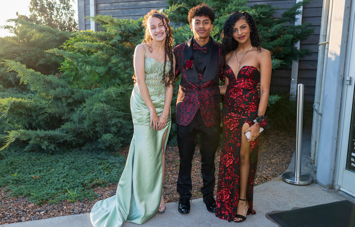 Three teens in formal gowns and suit posing together outdoors near a building exterior with greenery behind them, smiling at the camera.