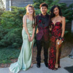 Three teens in formal gowns and suit posing together outdoors near a building exterior with greenery behind them, smiling at the camera.