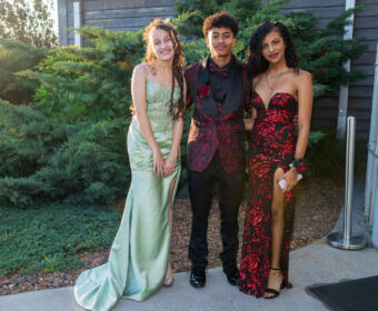 Three teens in formal gowns and suit posing together outdoors near a building exterior with greenery behind them, smiling at the camera.