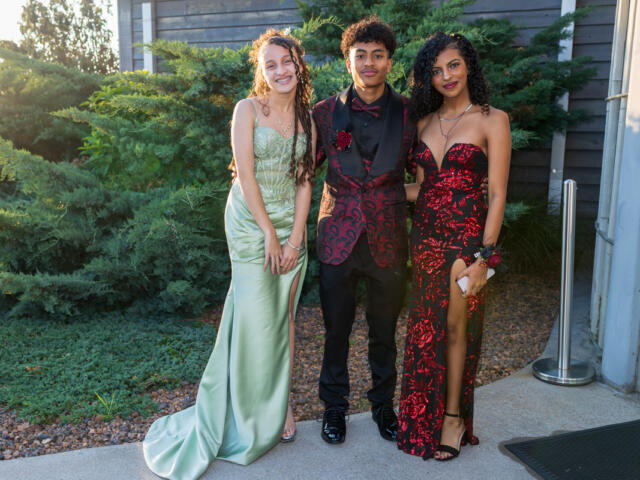 Three teens in formal gowns and suit posing together outdoors near a building exterior with greenery behind them, smiling at the camera.