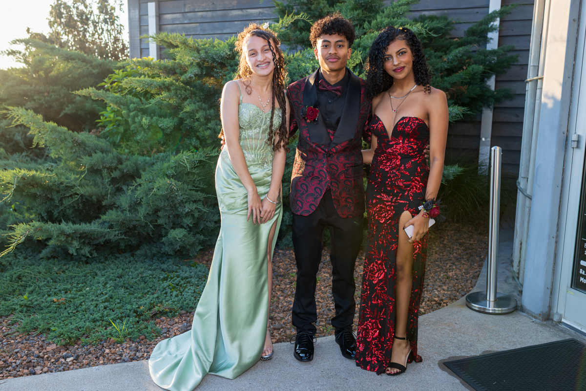 Three teens in formal gowns and suit posing together outdoors near a building exterior with greenery behind them, smiling at the camera.