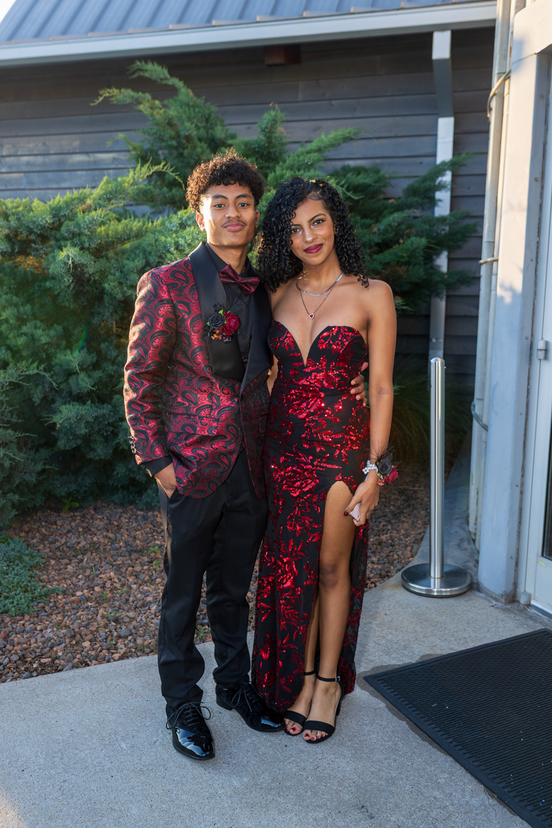 A young couple dressed for a formal event posing together outside a building; the man wears a red patterned suit with a black shirt and bow tie, and the woman wears a red and black sequined gown with a high slit.