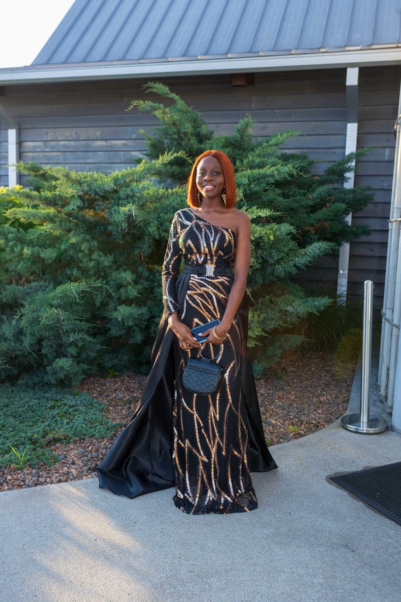 Woman in a one-shoulder black and gold sequined evening gown, holding a blue clutch and small purse, standing outdoors near shrubs.