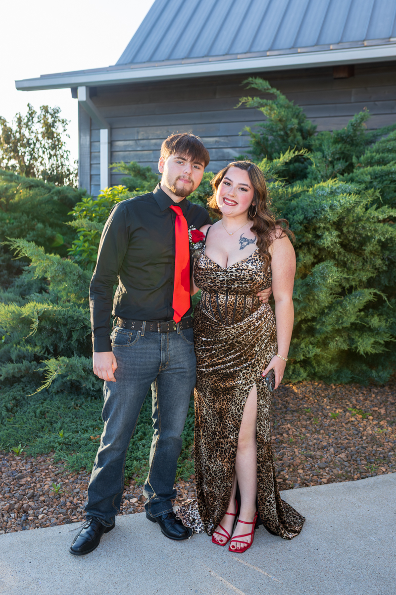 Young couple posing outdoors; woman in a leopard-print gown with red heels, man in a black shirt and red tie.