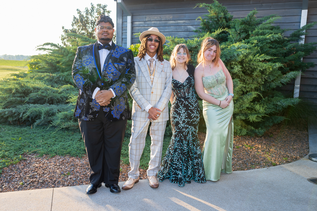 Four young adults in formal wear posing outdoors beside shrubs and a wooden building in sunlight.