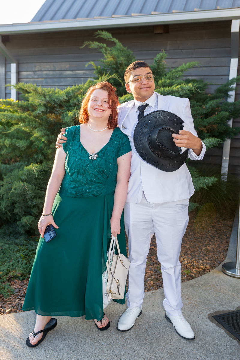 Smiling couple outdoors in formal attire: woman in a green lace dress with a pearl necklace, holding a beige handbag; man in a white suit holding a black hat beside her.